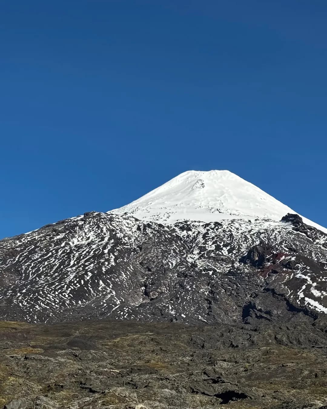 Volcán Villarrica con su cumbre nevada bajo un cielo azul despejado, desde el sendero de roca volcánica